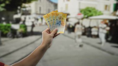 Açık bir şehir caddesinde bir adam tarafından tutulan İsviçre banknotları, hareketli şehir ortamında finans ve para birimi temalarını gözler önüne seriyor..