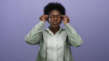 Yüzü kapalı bir kadın, mor arka plan karşısında stresli. Gözlüklü, yeşil ceketli, endişeli bir şekilde yukarı bakıyor..