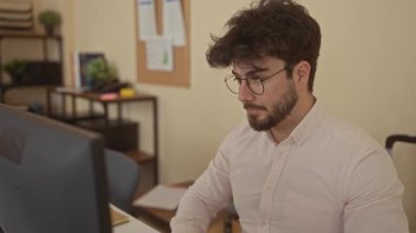 Gözlüklü genç adam ofis ortamında sessiz bir jest yapıyor. Profesyonel bir ortamda raflar ve klasörlerle çevrili bir masada odaklanma ve konsantrasyon gösteriyor..