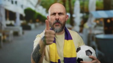 Sakallı bir adam futbol topuyla baş parmağını kaldırıyor, atkı takıyor, açık hava kafeleri olan canlı bir şehir sokağında duruyor, coşkuyu ve takım ruhunu yansıtıyor..