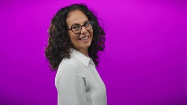 Kıvırcık saçlı ve gözlüklü olgun bir kadın kameraya gülümser ve izole edilmiş mor bir arkaplan karşısında onay verir, pozitiflik ve onay anı yakalar..