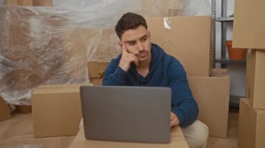 Etrafı kutularla çevrili yeni bir evdeki genç adam dizüstü bilgisayar kullanarak oturma odasında düşünce jesti yapıyor..