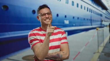 Gözlüklü ve dövmeli genç adam arka planda kırmızı ve beyaz çizgili gömlekli büyük bir yolcu gemisiyle bir limanda neşeyle gülümsüyor ve canlı bir sahil havası yaratıyor..