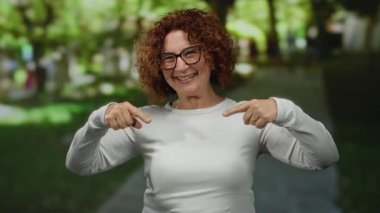 Güneşli bir parkta gülümseyen ve el kol hareketi yapan bir kadın, açık havada, gözlük takıyor ve günlük beyaz kıyafetler giyiyor, bulanık yeşillik ve ağaçlarla çevrili, neşeli bir anı yakalıyor..