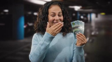 Otoparkın içinde orta yaşlı bir kadın Avustralya doları tutuyor, gülümsüyor ve onaylıyor, başarıyı ve pozitifliği finansal bağlamda iletiyor..