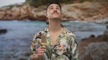 Genç İspanyol adam deniz kenarında dondurma yiyor, güneşli bir plaja bakıyor, açık deniz manzarasında kaygısız bir turist havası yaratıyor..