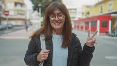Şehir sokaklarında gülümseyen son sınıf öğrencisi kadın canlı şehir zeminine karşı gözlük ve ceket takarak barış işareti yapıyor.