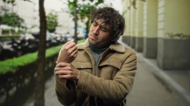 Bulutlu bir günde şehir sokaklarında acı içinde bileğini tutan genç bir adam açık hava ortamında rahatsızlık ve endişe taşıyor..