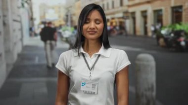 Uzun saçlı, beyaz tişörtlü genç bir kadın kalabalık bir sokak ortamında ellerini uzatarak açık hava şehir atmosferini gözler önüne seriyor..