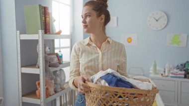 Çamaşır odasında çamaşır yıkayan bir kadın elinde sepet olan, çizgili tişört giyen, oyuncaklarla ve raflarla çevrili sıcak bir ev ortamında ev işleri yapan bir kadın..