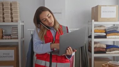 Kırmızı yelek giyen genç bir kadın ev içi bağışta dizüstü bilgisayar kullanıyor. Etrafı kutular ve kıyafetlerle çevrili.