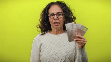 Orta yaşlı bir kadın, izole edilmiş sarı arka planda dolarları tutuyor. Parayla şaşkınlık ve heyecan yaratıyor..