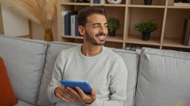 Elinde tabletle kanepede oturan genç bir adam, evin içinde gülümsüyor, modern bir oturma odasında rahat bir ortam ile ev dekoruyla çevrili..