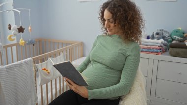 Hamile kadın kitap okuyor. Yatak odasında beşiği ve oyuncaklarıyla oturuyor. Sakin, rahat ve kapalı bir ortam sergiliyor..