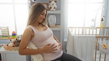 Yatak odasında yatakta oturan bir kadın, evde rahat bir şekilde hamile karnına gülümseyen bir bebek beşiği ile ev ortamında sıcaklığı ve beklentiyi sembolize ediyor..
