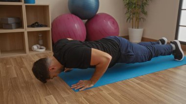 Modern spor salonunda spor aletleri ve denge toplarıyla çevrili mavi minder üzerinde şınav çeken atletik bir adam.