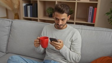 Akıllı telefonu ve kırmızı kupası olan genç bir adam modern bir dairenin oturma odasında bir kanepede oturuyor, rahatlamış ve ekrana odaklanmış, etrafı kitaplar ve dekorlarla çevrili..