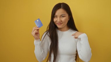 Kadının elinde mavi bir kredi kartı var. Sarı arka planda gülümsüyor. Beyaz süveter giyiyor. Finans konseptini gösteriyor..