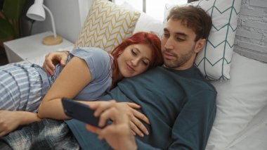 Bir çift yatak odasında dinlenirken, kadın kafasını erkeğin göğsüne yaslarken, o da akıllı bir telefon tutuyor, rahat bir evde samimiyet ve rahatlama tasvir ediyor..