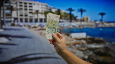 Sahilde bir sokakta tek başına dolar tutan bir adam, arka planda açık mavi deniz ve palmiye ağaçları olan canlı bir yaz tatili atmosferini yakalıyor..