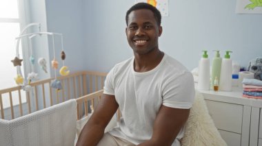 Sıcak bir yatak odasında gülümseyen genç bir adam, bebek beşiğinin yanında oturuyor ve evde rahat bir ortam yaratıyor..