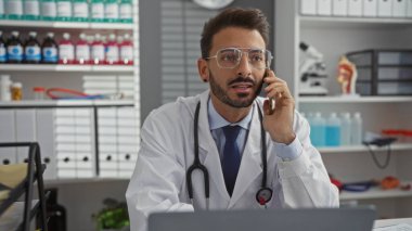 Tıbbi malzemeler ve mikroskopla çevrili, boynunda steteskopla klinik odasında konuşan İspanyol erkek doktor..