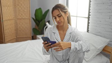 Kadın rahat yatak odasında pijama, sabah ışığı, kapalı ev, beyaz tuğla duvar, barışçıl, bitkiler, modern dekor, rattan mobilya ile akıllı telefon kullanarak yatakta oturuyor..