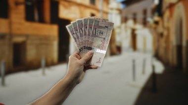 Bir şehir caddesinde Suudi Arabistan riyallerini elinde tutan adam, açık bir şehir ortamında finans ve uluslararası ekonomi üzerine açık bir odak noktasıyla para gösteriyor..