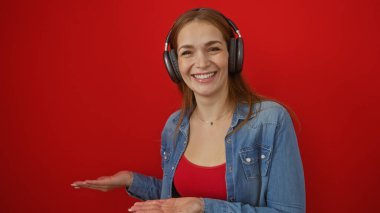 Kırmızı arka planda kulaklık takan, geniş avuç içi ile gülümseyen kadın, canlı ortama zıtlık ekleyen kot gömlek, yaratıcı ve anlamlı medya projeleri için mükemmel..