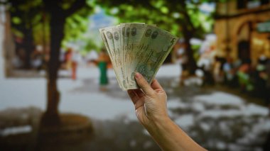 Kentsel sokak ortamında elinde Dirham banknotları tutan adam, ağaçların altında canlı şehir ortamında iş ve döviz alışverişini ön plana çıkarıyor..