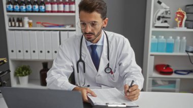 Tıbbi ekipman ve belgelerle çevrili, gözlüklü, stetoskop takan, odaklanmış ve profesyonel bir şekilde klinik odasında çalışan genç bir erkek doktor..