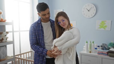 Bebek dekoru ve pencerenin doğal ışığıyla çevrili bir şekilde battaniyeye sarılı yeni doğmuş bebeği kucaklayan çift..