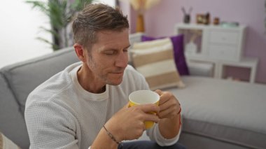 Yakışıklı bir adam, rahat oturma odasında kahve içiyor modern dekorlar ve yumuşak mobilyalarla, huzurlu ve rahat bir ev atmosferi yakalıyor..