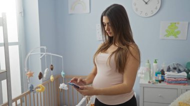 Oyuncak ve bebek malzemeleriyle çevrili, rahat bir bebek odasında akıllı telefon kullanan bir kadın, ev içi yatak odası ortamında, modern bir aile yaşam tarzını aktarıyor..