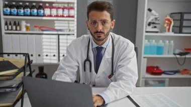 Bir klinik odasında, gözlük ve laboratuvar önlüğü takmış, etrafı tıbbi malzeme ve ekipmanlarla çevrili, profesyonellik taslayan genç bir İspanyol erkek doktor..