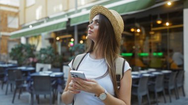 Esmer saçlı ve hasır şapkalı bir kadın, kafenin yakınındaki canlı bir şehir sokağını keşfetmekten zevk alıyor. Elinde akıllı bir telefonla, günlük bir kıyafet ve sırt çantası takıyor..