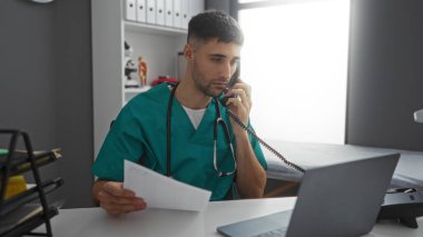 Parlak klinik odasında steteskopla konuşan, tıbbi önlüklü genç adam, belgeler ve laptopla çalışıyor, sağlık hizmetleri ayarları yapıyor..