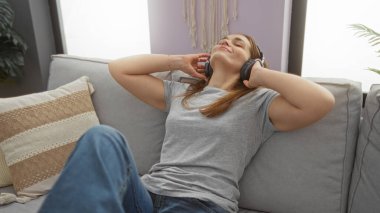 Kanepede dinlenen, rahat bir oturma odasında kulaklıkla müzik dinleyen, rahat bir anın tadını çıkaran, günlük kıyafetleri ve doğal ışığı olan bir kadın..