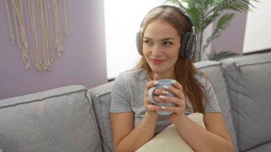 Modern oturma odasında kulaklıklarla dinlenen bir kadın elinde bir kupayla evin içinde müzikten zevk alan genç ve çekici bir yaşam tarzını yansıtan rahat bir atmosferle..