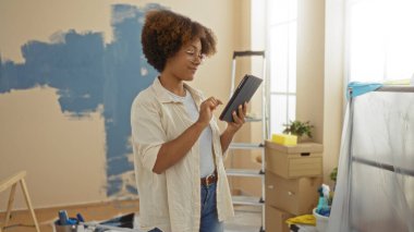 Tablet kullanan bir kadın yeni boyanmış oturma odasında kutular ve merdiven ile ev iyileştirme projesini gösteriyor..
