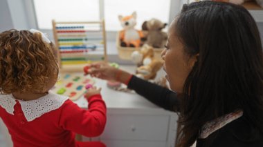 Anaokulunda kızıyla oynayan bir kadın oyuncaklarla dolu, aile sevgisini ve birlikteliğini gösteren, bağ ve eğitici oyunu ön plana çıkaran bir kadın..