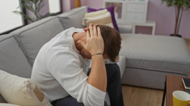Genç adam, modern oturma odasındaki koltukta oturmuş sıkıntılı bir şekilde hayata dair zorlukları yansıtan stresi anlatıyor.