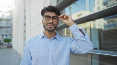 Sakallı ve gözlüklü genç adam şehir sokaklarında güler yüzlü modern binalarla şehir yaşamını yansıtıyor..