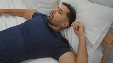 Latin kökenli genç bir adam rahat bir yatak odasında huzur içinde dinleniyor. Rahat ve rahat bir ortam ve günlük kıyafetlerle rahatlığı ön plana çıkarıyor.