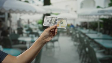 Şehir caddesindeki açık hava restoranının terasında elinde Amerikan Doları banknotuyla gezen adam şehir çevresiyle sıradan bir yemek sahnesi sergiliyor..
