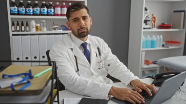 Hastane odasında çalışan bir doktor, labaratuar önlüklü İspanyol bir adam masasında steteskop, masada tıbbi malzemeler ve raflarda görünen belgeler..