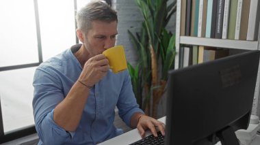 Modern ofiste kitaplık geçmişi olan bilgisayarda çalışırken kahve içen genç bir adam.