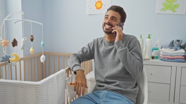 Bebek odasında beşik ve oyuncaklarla telefonda konuşurken gülümseyen genç İspanyol adam, rahat ev ortamının keyfini çıkarıyor..