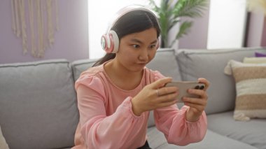 Modern oturma odasında akıllı telefondan kulaklık takan, odaklanmış ve teknolojiyle meşgul bir kadın..