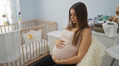 Bebek odasında rahat bir sandalyede oturan kadın, pelüş oyuncaklarla dolu ahşap bir beşiğin yanında hamile karnına dokunuyor, beklenti ve annelik duyguları uyandırıyor..